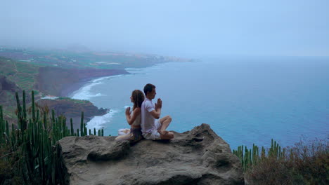 Amidst-mountain-views-and-the-vast-ocean,-a-man-and-woman-sit-back-to-back-on-a-rock,-immersing-themselves-in-meditation-and-yoga